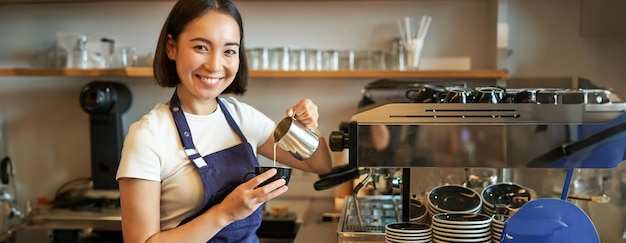 Photo gratuite une barista asiatique souriante fait du café, verse du lait cuit à la vapeur dans un cappuccino, fait du latte dans une tasse.