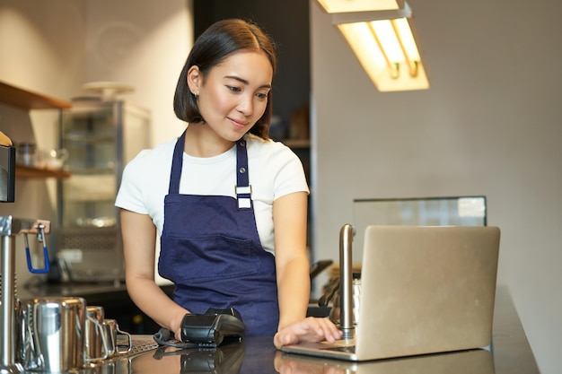 Barista asiatique souriante travaillant dans un café servant un client regardant un ordinateur portable tout en utilisant un lecteur de carte