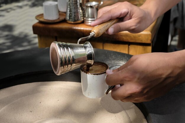 Barista verse du café turc fraîchement préparé dans une tasse en gros plan Un barista professionnel prépare le café à la main de manière traditionnelle dans le sable Espace de mise au point sélective pour le texte