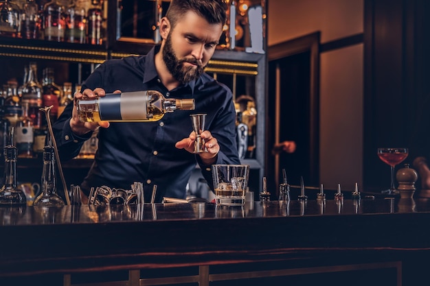 Un barman brutal élégant dans une chemise noire prépare un cocktail au fond du comptoir du bar.