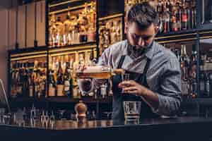 Photo gratuite barman brutal élégant dans une chemise et un tablier fait un cocktail au comptoir du bar.