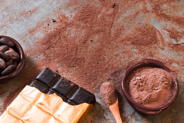 Barre de chocolat noir avec poudre de cacao dispersée et bol de haricots sur fond rustique