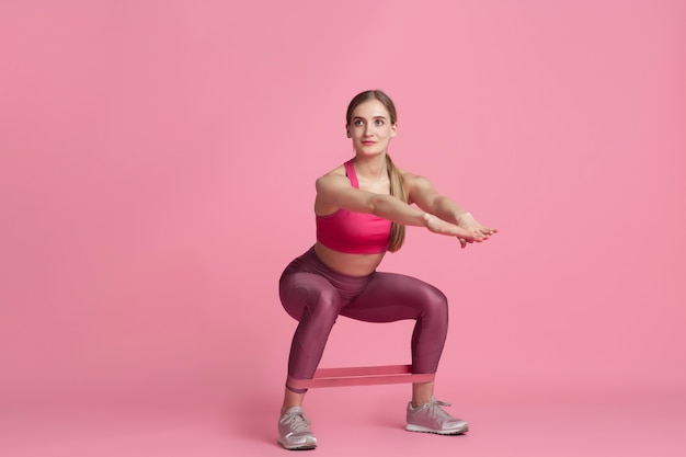 Bas du corps. Belle jeune athlète féminine pratiquant, portrait rose monochrome. Modèle caucasien de coupe sportive avec élastiques. Musculation, mode de vie sain, concept de beauté et d'action.