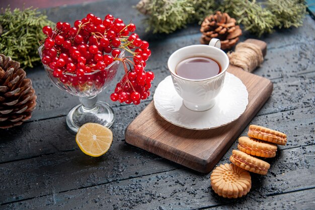 En bas vue rapprochée de groseille rouge dans un verre une tasse de thé sur une planche à découper tranche de pommes de pin au citron et biscuits sur table en bois foncé