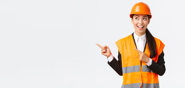 Bâtiment, construction et concept industriel. Architecte asiatique souriant, enthousiaste et intéressé, ingénieur en casque et vêtements de sécurité à la recherche, pointant le coin supérieur gauche vers un projet intéressant