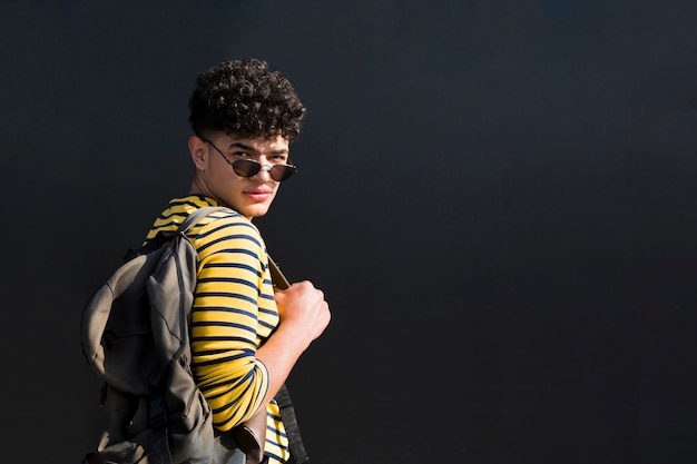 Photo gratuite beau adolescent ethnique à lunettes de soleil et chemise rayée avec sac à dos, regardant en arrière