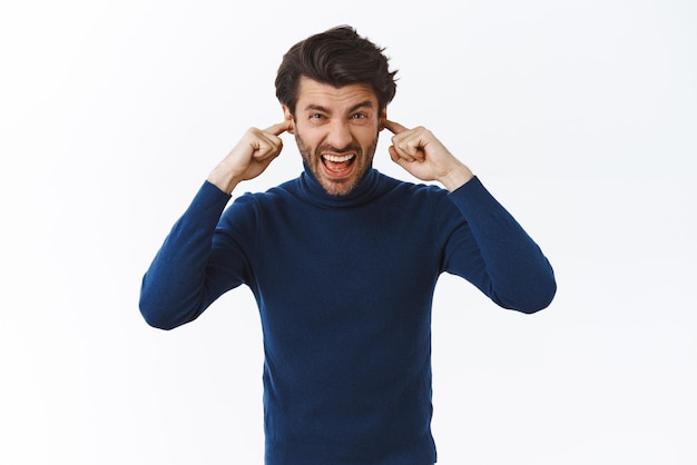 Photo gratuite un beau barbu dérangé ferme les oreilles avec les doigts et crie en plissant les yeux mécontent de se sentir mal à l'aise d'entendre un bruit fort et ennuyeux fatigué des voisins ayant des travaux de réparation