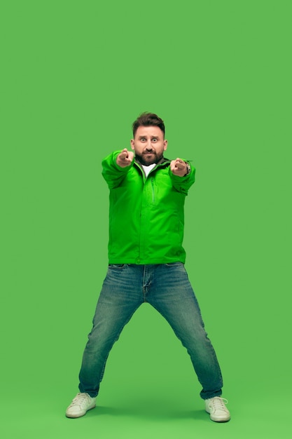 Beau barbu souriant heureux jeune homme regardant et pointant la caméra isolée sur studio vert branché vif.