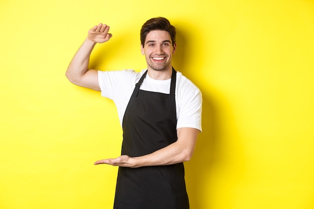 Beau barista souriant montrant quelque chose de long ou de grand, debout sur fond jaune