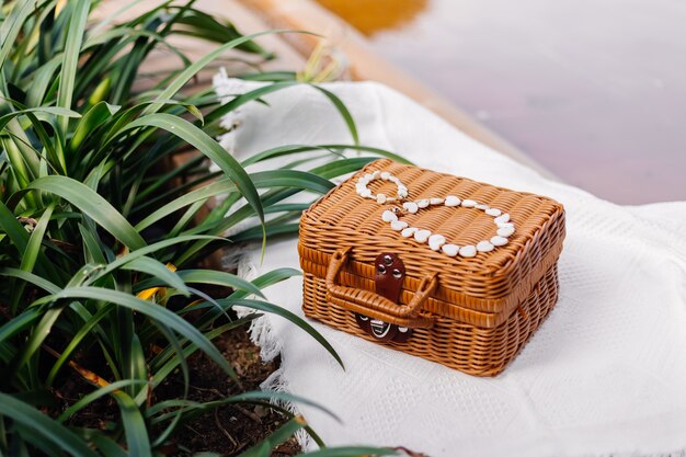 Beau bracelet et collier de bijoux en coquillage blanc sur une poitrine en osier marron et un tapis de plage blanc par Tropical Leafs Outdoor Shot