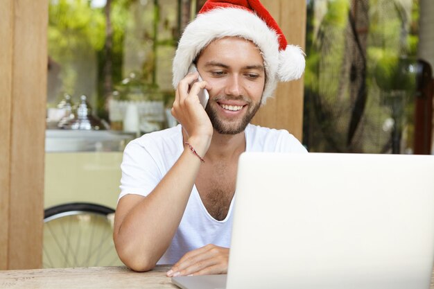 Beau caucasien au chapeau de père Noël, parler au téléphone mobile, assis devant un ordinateur portable ouvert, souriant, ayant une expression de visage joyeuse, profitant des vacances du nouvel an dans un pays chaud