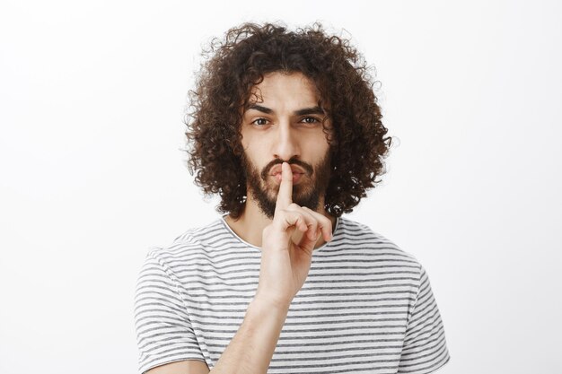 Beau collègue masculin confiant dans un t-shirt rayé élégant, faisant un geste chut, disant chut avec l'index sur la bouche, fronçant les sourcils, étant ludique et concentré