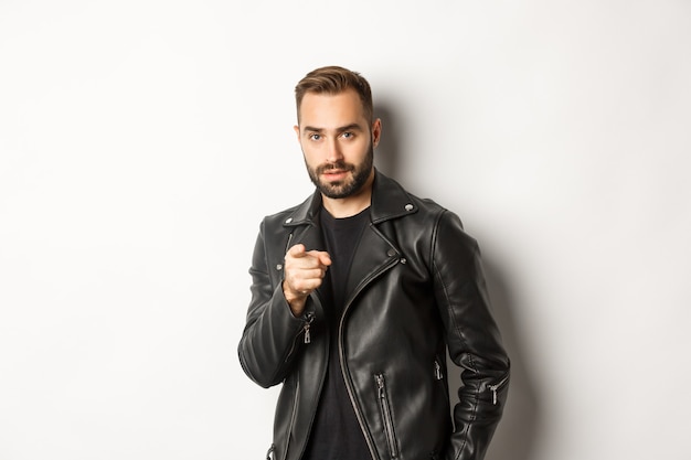 Beau et cool mec barbu pointant du doigt la caméra, vêtu d'une veste en cuir, debout impertinent