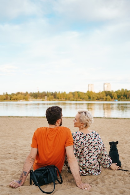 Beau couple de détente sur le sable étreignant et s'embrassant tout en jouant avec leur chien positif