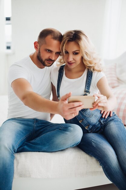 Beau couple de femme enceinte et son mari portant des jeans assis s'embrassant et prenant l'autoportrait Jeune maman et papa tenant un téléphone intelligent et prenant une photo