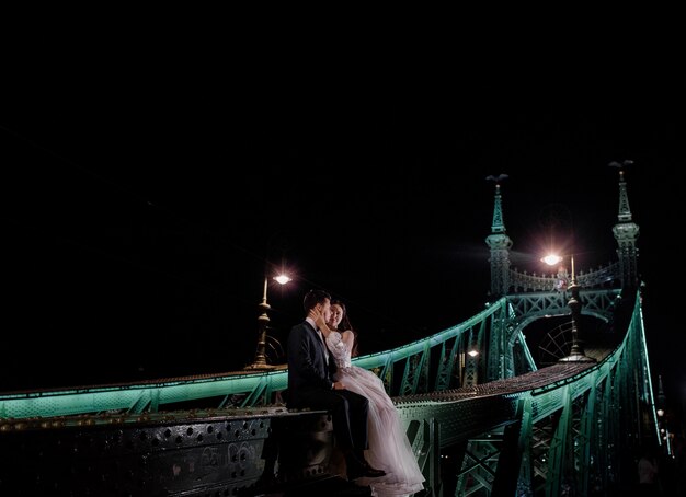 Beau couple de mariage est assis sur le pont illuminé dans la nuit noire et s'embrasse