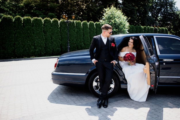 Beau couple de mariage sourit dans la voiture noire sur la journée ensoleillée, vêtu de tenues de mariage élégantes avec bouquet rouge