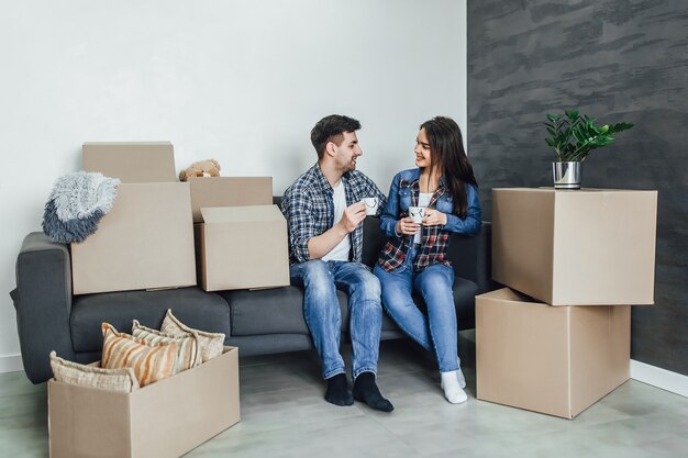 Un beau couple en vêtements décontractés discute du plan de leur nouvelle maison et sourit en étant allongé sur le canapé près des boîtes de déménagement