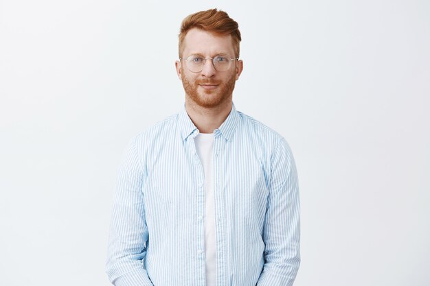 Beau entrepreneur homme rousse créatif et intelligent debout dans une chemise décontractée et des lunettes sur un mur gris, souriant amical, regardant avec un regard réfléchi et déterminé