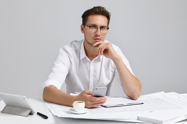 Beau entrepreneur masculin barbu assis au bureau dans l'intérieur de bureau moderne à l'aide d'un téléphone portable tout en étudiant le plan, ayant un regard sérieux réfléchi, touchant le menton. Personnes, travail et profession