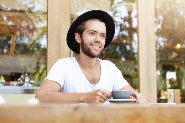 Beau étudiant masculin à la mode avec une barbe épaisse assis à une table en bois avec une tasse et boire du café, ayant une expression de visage joyeuse et joyeuse