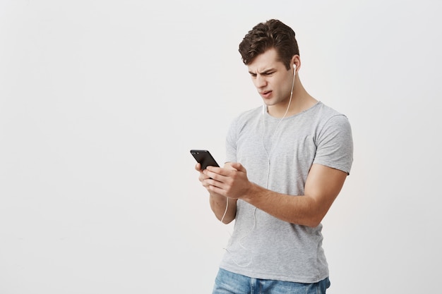 Beau étudiant masculin tient un téléphone portable dans ses mains, regarde avec une expression confuse à l'écran, porte des écouteurs blancs, fronce les sourcils. Un homme de race blanche confondu avec le message qu'il a reçu de sa petite amie.