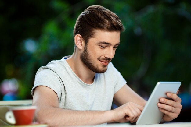 Beau gai jeune homme souriant assis à la table dans un café en plein air tenant la tablette à l'écran.