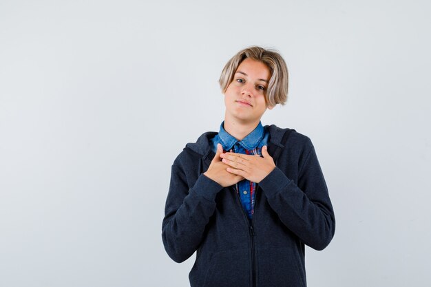 Beau garçon adolescent avec les mains sur la poitrine en chemise, sweat à capuche et l'air reconnaissant, vue de face.