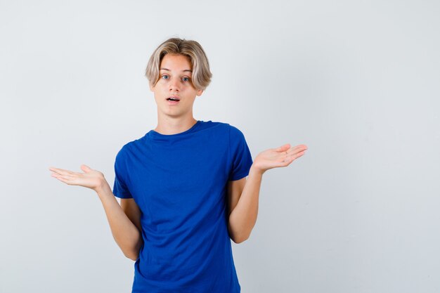 Beau garçon adolescent en t-shirt bleu montrant un geste impuissant et l'air perplexe, vue de face.