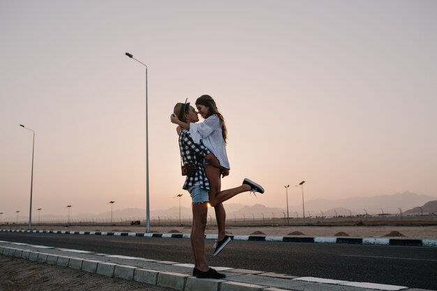 Beau garçon élégant au chapeau à la mode tenant sa petite amie mince en chemisier blanc, debout sur la route en soirée d'été. Beau jeune couple étreignant à une date avec coucher de soleil incroyable