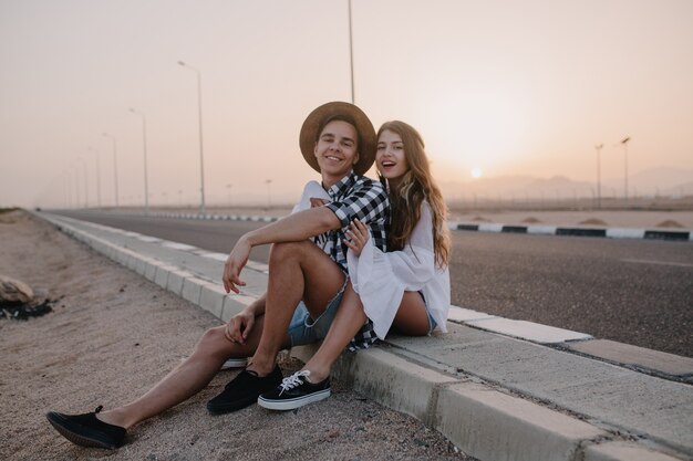 Beau garçon portant chapeau et femme en chemisier vintage blanc assis ensemble sur la route et jouit de beau coucher de soleil. Charmante jeune femme aux cheveux longs au repos près de l'autoroute avec son petit ami