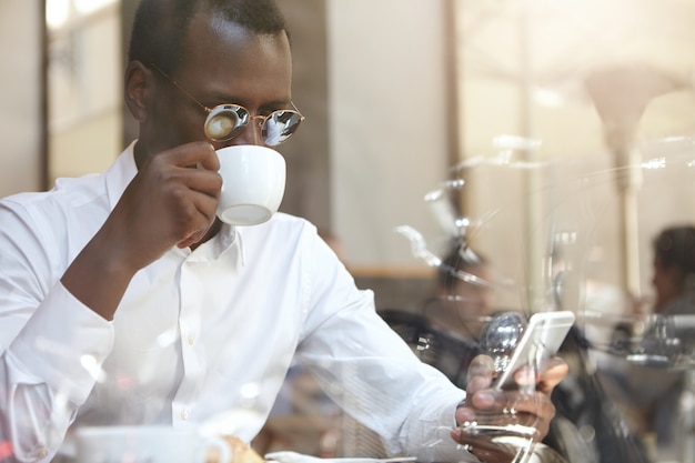 Beau gestionnaire noir en tenue de soirée vérifiant le courrier électronique ou lisant les nouvelles du monde sur un téléphone portable numérique, buvant un cappuccino du matin, assis à table au café. Technologie, connexion et communication