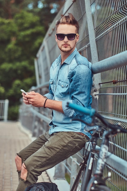 Photo gratuite beau hipster avec une coupe de cheveux élégante dans des lunettes de soleil se reposant après avoir fait du vélo, tient un smartphone.