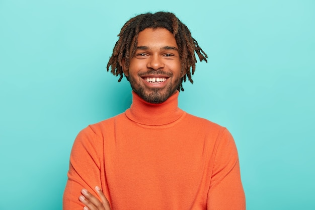 Beau hipster avec des dreads a un sourire agréable, a des dents blanches, heureux d'entendre de bonnes nouvelles, porte des vêtements lumineux