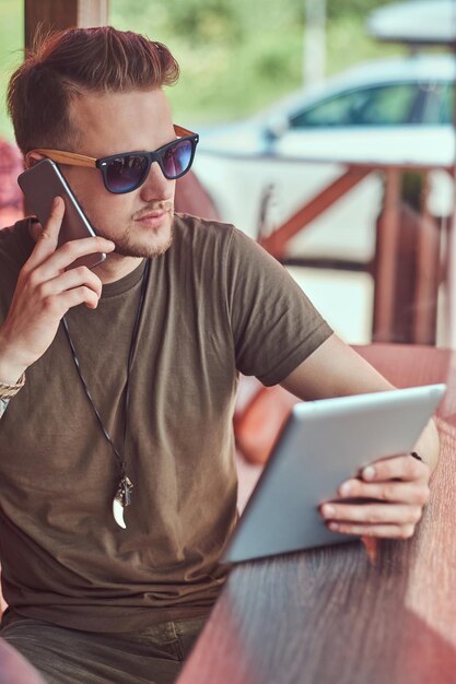 Beau hipster élégant est assis à une table dans un café en bordure de route, parle au téléphone et tient la tablette.