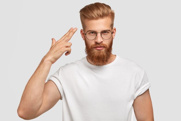 Beau hipster masculin avec barbe de gingembre et chaume, vêtu d'un t-shirt blanc décontracté, fait un geste de suicide, tire dans le temple, se sent fatigué des problèmes et de la vie difficile, isolé sur le mur