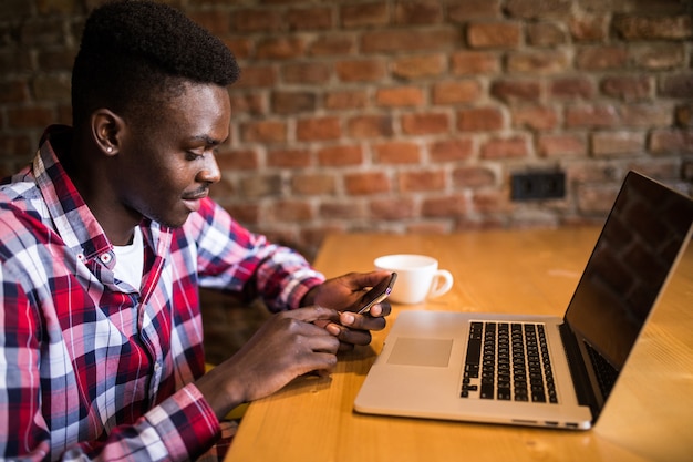 Beau jeune afro-américain souriant joyeusement en lisant des sms sur un téléphone intelligent, la messagerie à l'aide du wi-fi gratuit, prendre un café au café