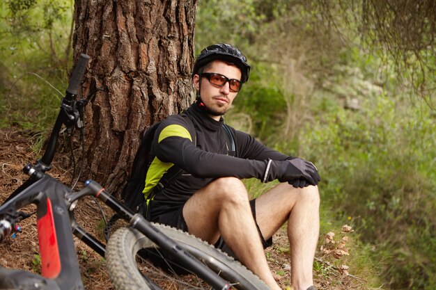 Beau jeune cavalier professionnel portant des lunettes et un casque assis sous un arbre, se détendre et admirer la belle vue après l'entraînement à vélo du matin sur un vélo d'appoint à moteur le week-end