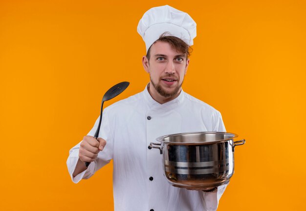 Un beau jeune chef barbu homme en uniforme blanc tenant une casserole avec une cuillère à fente tout en regardant sur un mur orange