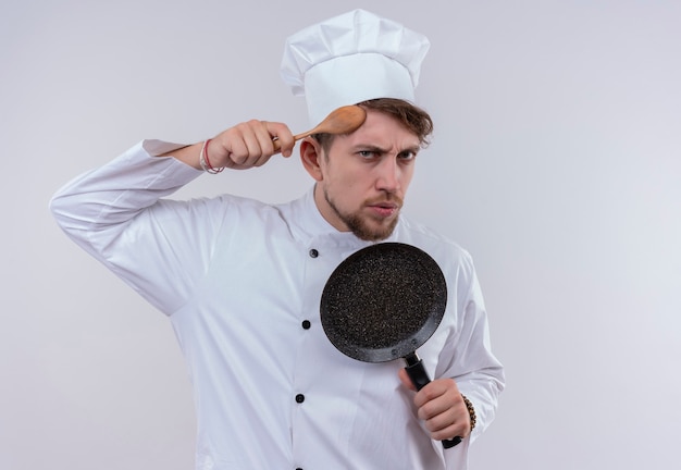 Un beau jeune chef barbu homme vêtu d'un uniforme de cuisinière blanche et chapeau montrant une poêle à frire tout en tenant une cuillère en bois sur sa tête sur un mur blanc