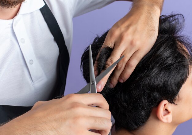 Beau jeune coiffeur en uniforme faisant la coupe de cheveux pour jeune client isolé sur fond violet