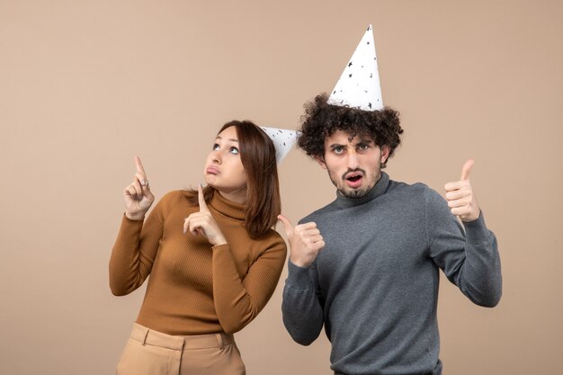 Beau jeune couple excité porte chapeau de nouvel an fille regardant au-dessus et debout près de gars sur l'image grise