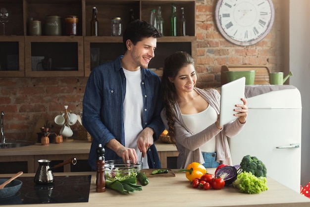 Un beau jeune couple utilise une tablette numérique et lit une recette sur un site web culinaire tout en cuisinant dans une cuisine loft à la maison, espace de copie