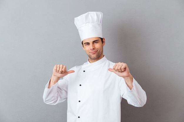 Beau jeune cuisinier en uniforme pointant vers lui.