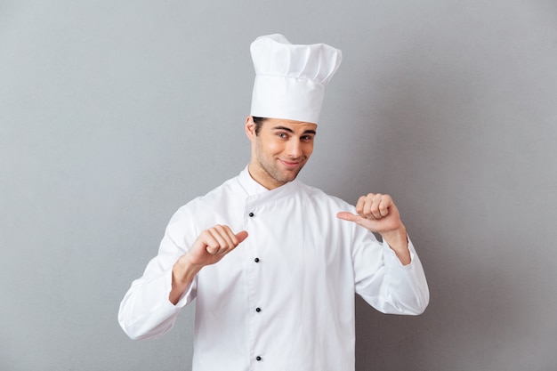 Beau jeune cuisinier en uniforme pointant vers lui.