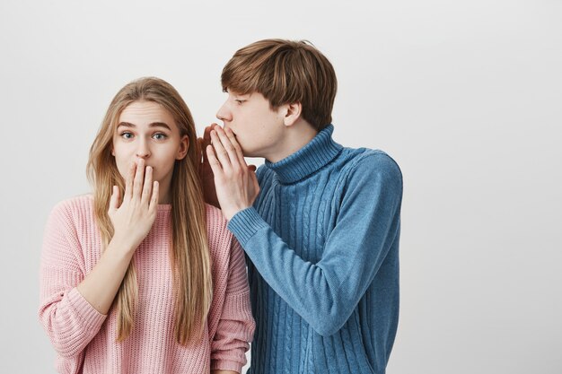 Beau jeune étudiant blond en pull bleu, chuchotant quelque chose à l'oreille d'une fille blonde élégante