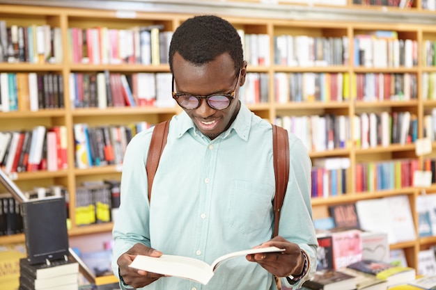 Beau jeune hipster afro-américain dans les tons tenant un livre ouvert dans ses mains, lisant son poème préféré, cherchant l'inspiration dans une bibliothèque publique ou une librairie. Gens, style de vie et loisirs