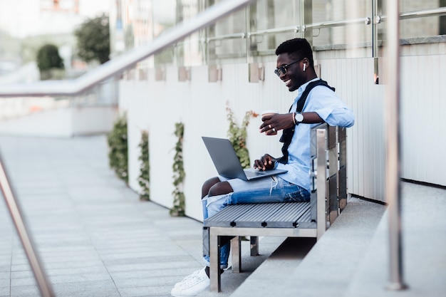 Un beau jeune homme d'affaires assis sur un banc avec son ordinateur portable dans une rue ensoleillée à côté d'un parc. Avec une tasse de café.