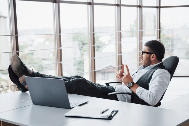 Beau jeune homme d'affaires avec des lunettes tenant ses jambes sur la table en regardant un ordinateur portable au bureau