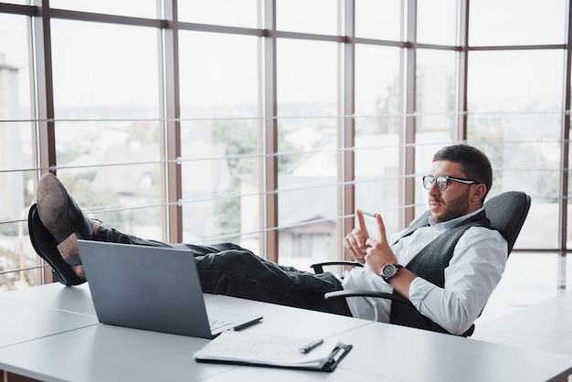 Beau jeune homme d'affaires avec des lunettes tenant ses jambes sur la table en regardant un ordinateur portable au bureau